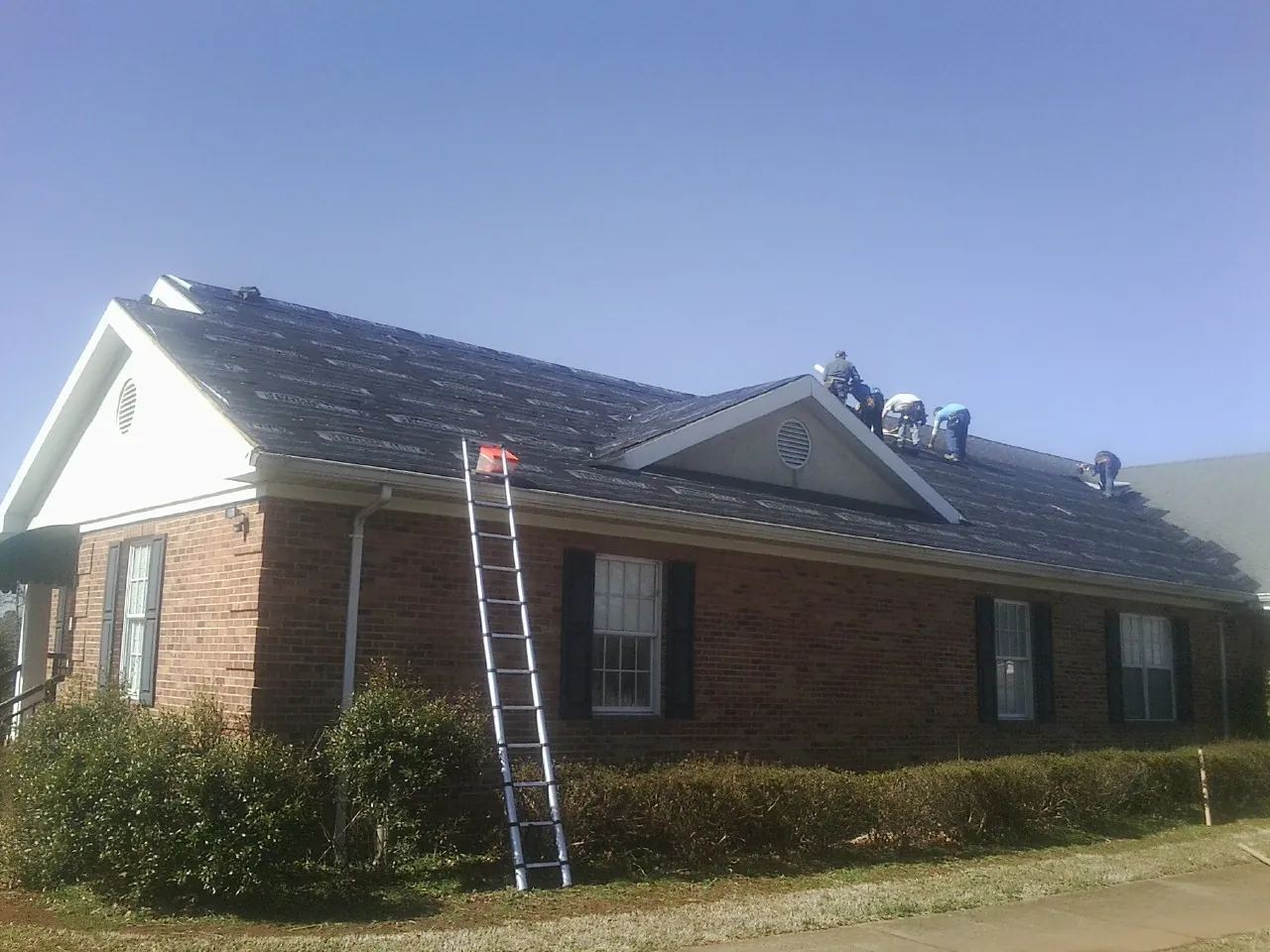Crew tearing shingles off a brick ranch roof with a ladder set — Emergency Roof Repair work by our crew in Sophia