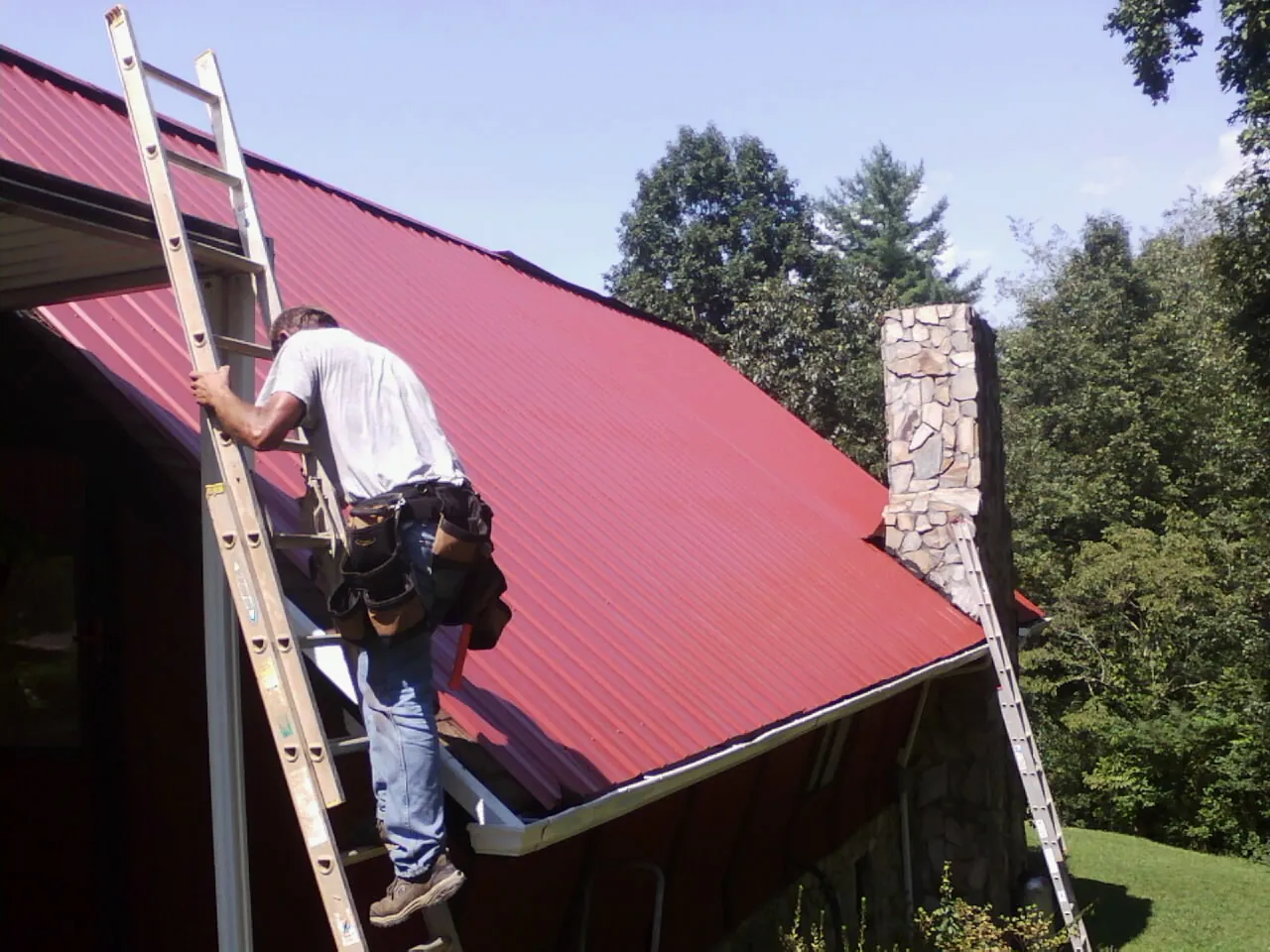 Roofer on a ladder beside a red metal roof and stone chimney — Soffit & Fascia Repair by Roofing Brothers in Sophia